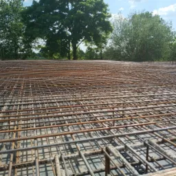 Siatka zbrojeniowa na betonowej płycie, widok z dołu pod kątem, z zielonymi drzewami w tle pod błękitnym niebem.