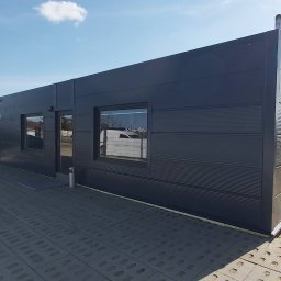 KONSTRUKT Grzegorz Czarnecki - Nowoczesna, grafitowa hala z płyt warstwowych, z widocznymi oknami i wejściem, na betonowej nawierzchni z otworami, pod błękitnym niebem z pojedynczą chmurą.