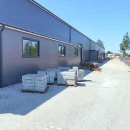 KONSTRUKT Grzegorz Czarnecki - Gotowe bloki betonowe na paletach przed budynkiem hali z szarymi panelami, ułożone na kostce brukowej w słoneczny dzień.