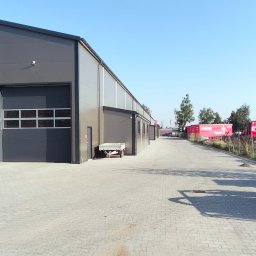 KONSTRUKT Grzegorz Czarnecki - Nowoczesna hala magazynowa z szarej płyty warstwowej, widok z zewnątrz na plac z kostki brukowej i zaparkowane czerwone ciężarówki w tle.