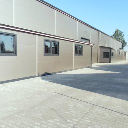 KONSTRUKT Grzegorz Czarnecki - Nowoczesna hala przemysłowa z paneli warstwowych w odcieniach szarości, z widocznymi oknami i brukowaną nawierzchnią przed budynkiem w słoneczny dzień.