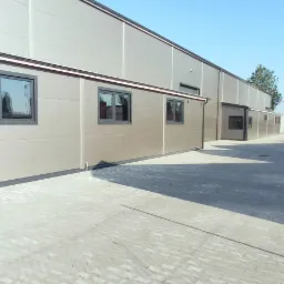 Nowoczesna hala przemysłowa z paneli warstwowych w odcieniach szarości, z widocznymi oknami i brukowaną nawierzchnią przed budynkiem w słoneczny dzień.