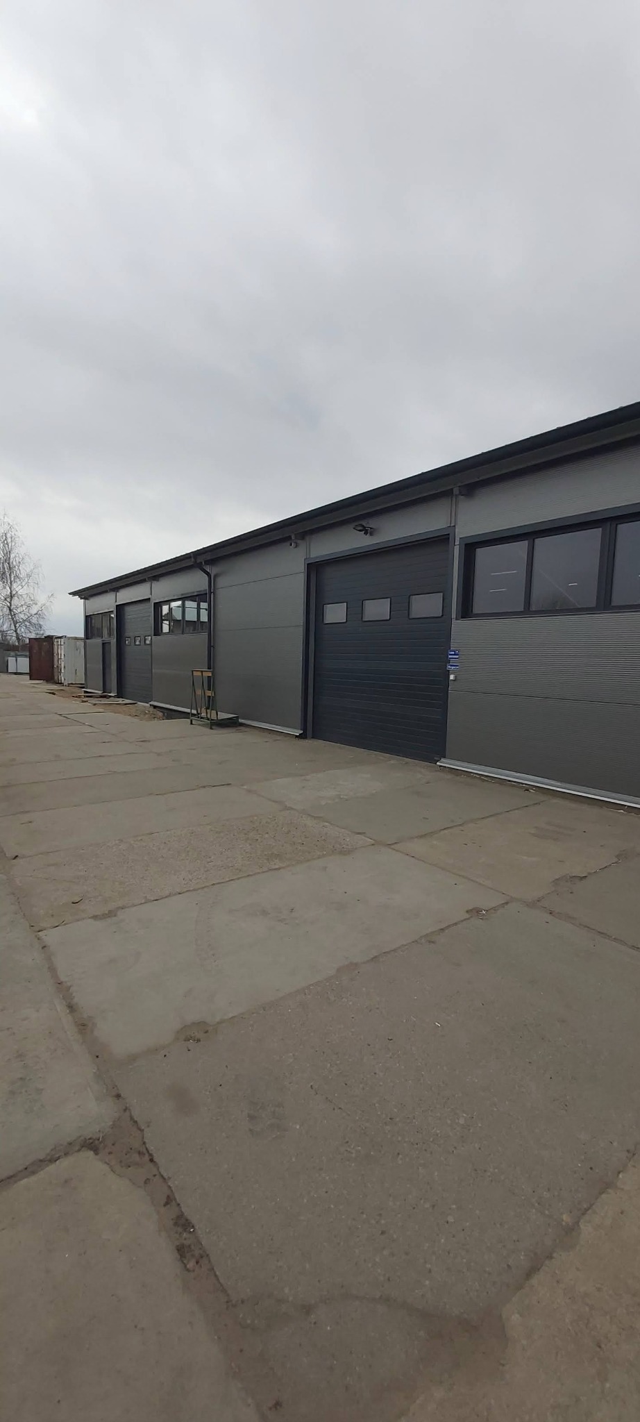 Zewnętrzny widok hali przemysłowej z szarymi ścianami z paneli warstwowych i segmentowymi bramami garażowymi, betonowy plac przed budynkiem, pochmurne niebo.