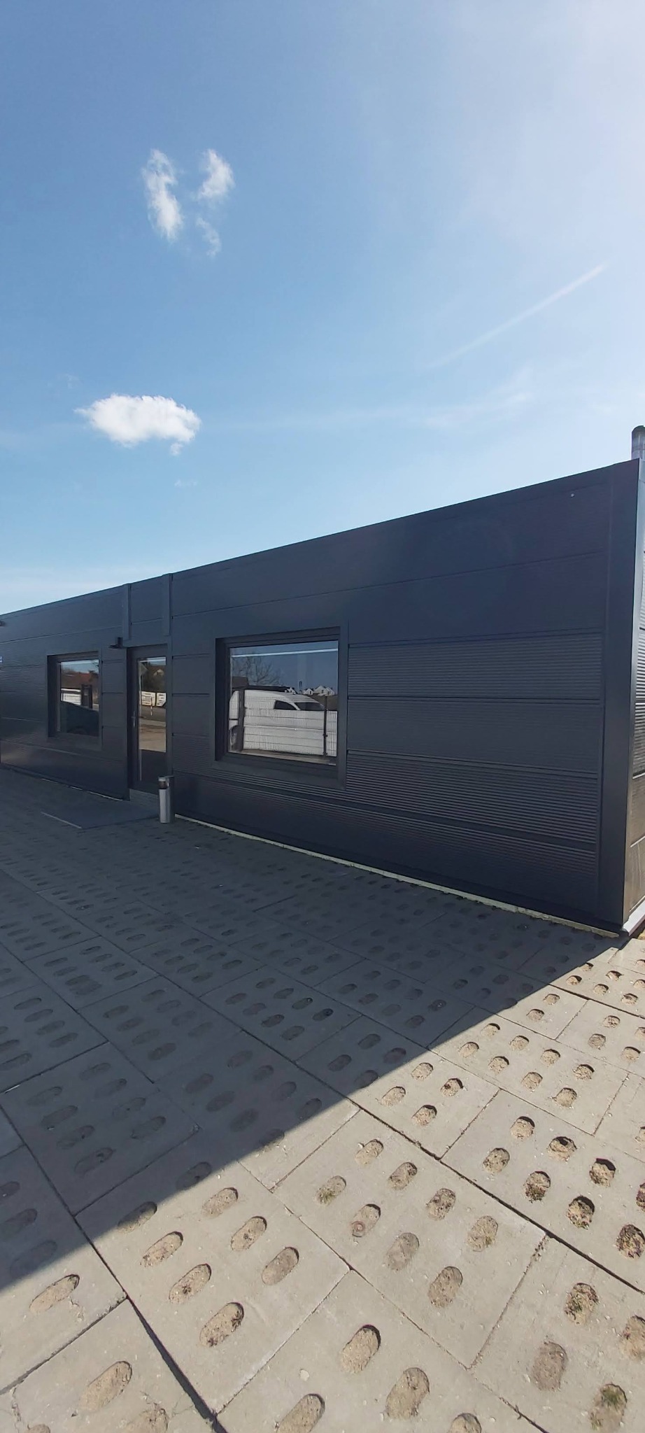 Nowoczesna, grafitowa hala z płyt warstwowych, z widocznymi oknami i wejściem, na betonowej nawierzchni z otworami, pod błękitnym niebem z pojedynczą chmurą.