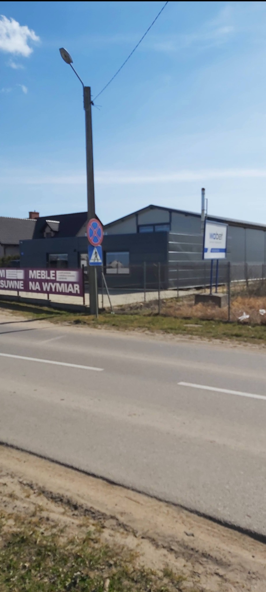 Nowoczesna hala przemysłowa z szarej płyty warstwowej, widoczna zza drogi, z tablicą reklamową firmy budowlanej i znakami drogowymi.