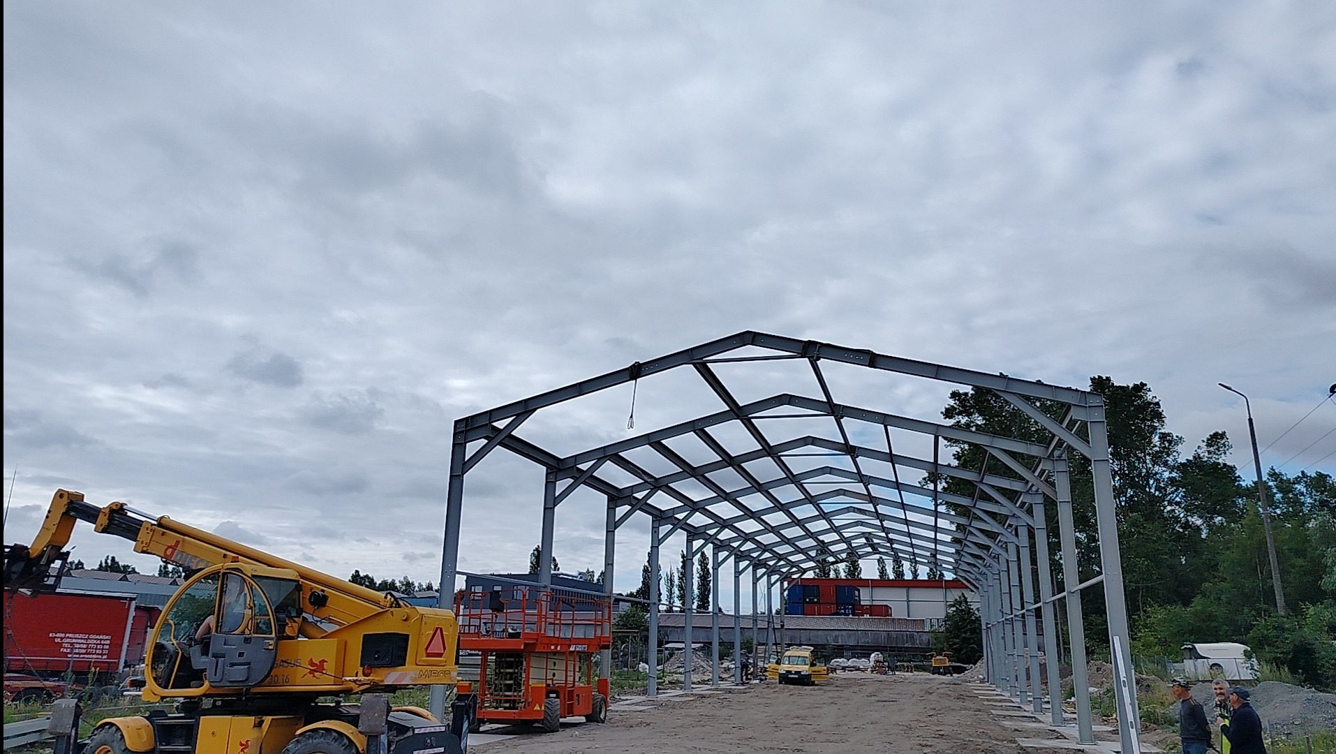 Wznoszenie stalowej konstrukcji hali na tle pochmurnego nieba, widoczne podnośniki i pracownicy budowlani.