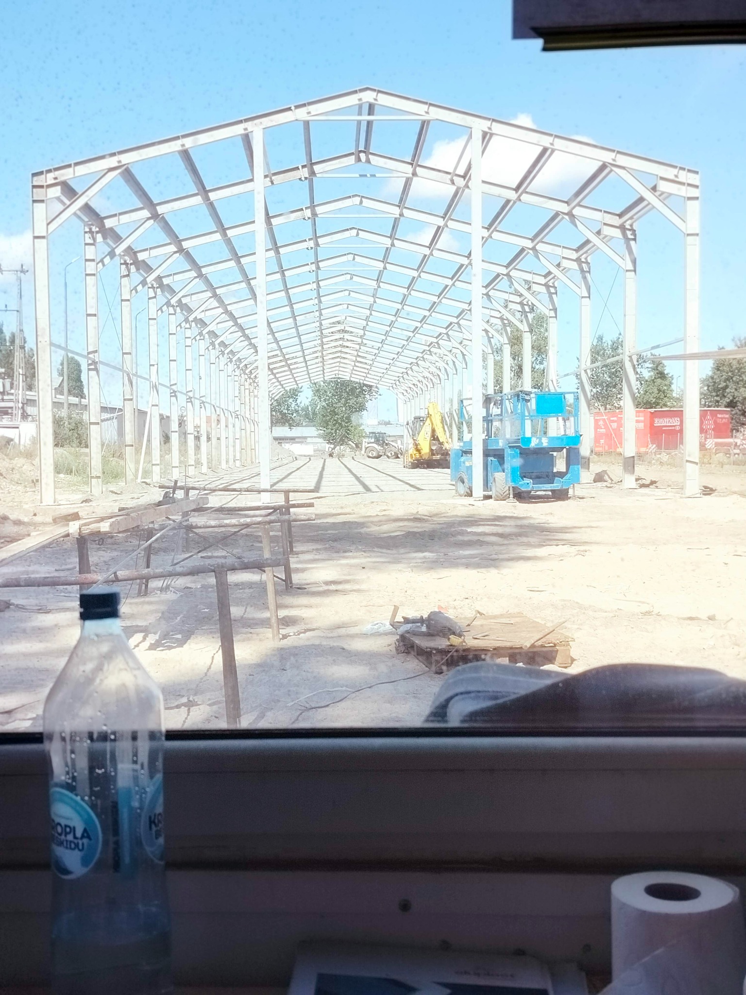 Wnętrze pojazdu z widokiem na plac budowy hali stalowej w trakcie montażu, z widocznym szkieletem konstrukcji, sprzętem budowlanym i materiałami.