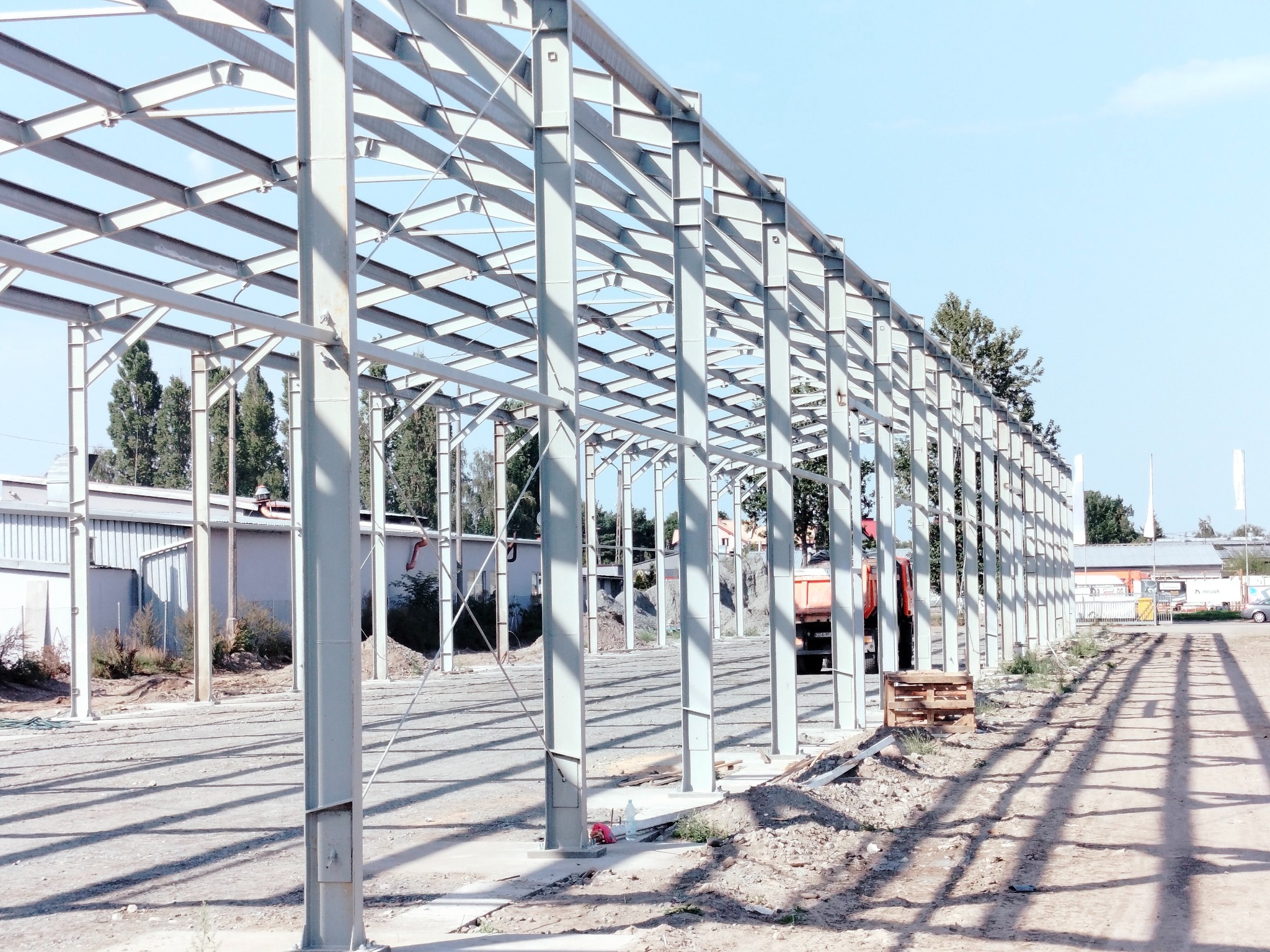 Konstrukcja stalowa hali w trakcie budowy, widoczne elementy szkieletu, w tle budynek magazynowy i ciężarówka
