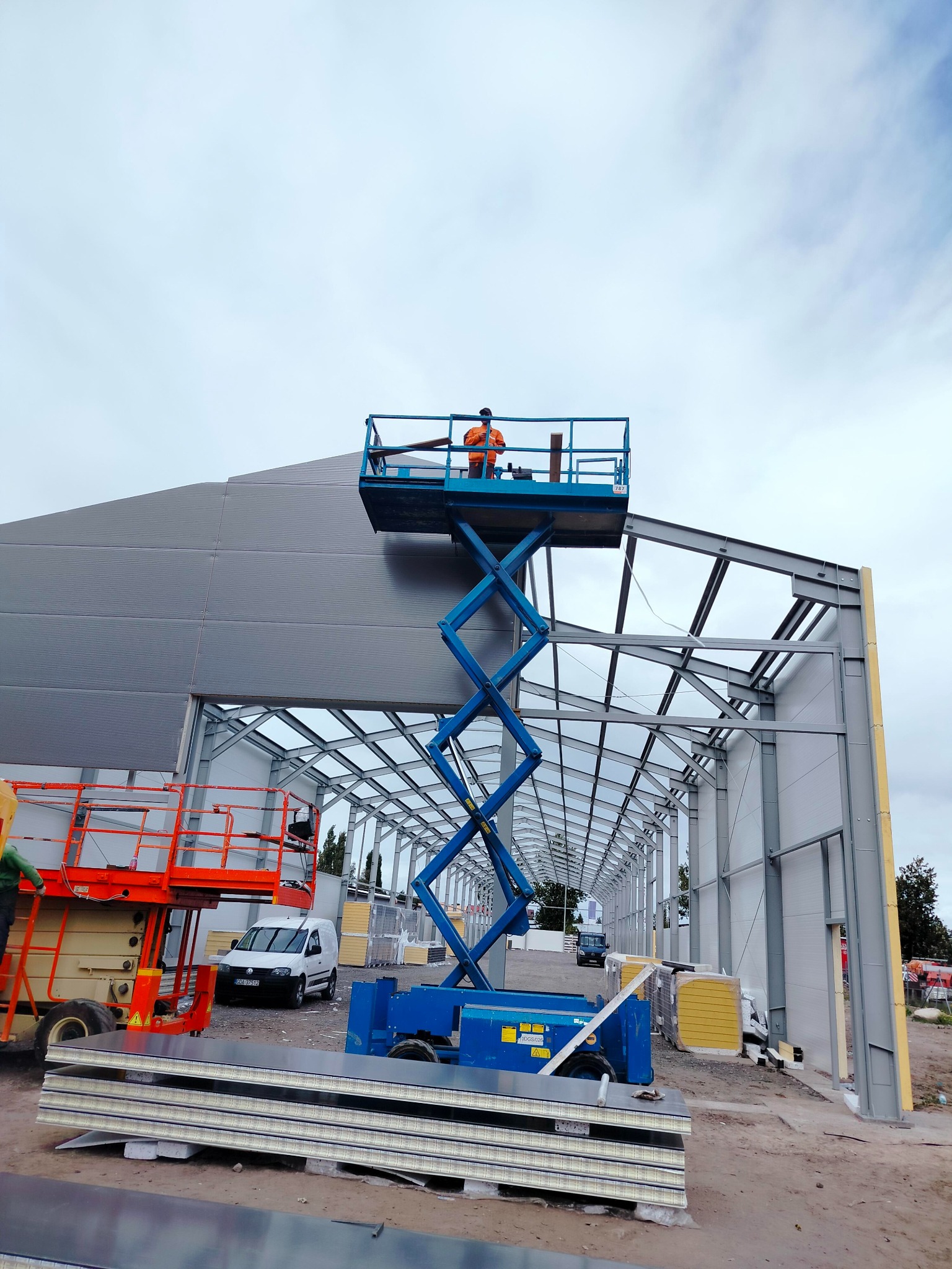 Montaż szarych płyt warstwowych na stalowej konstrukcji hali z użyciem podnośnika nożycowego; w tle widoczny inny podnośnik i samochód dostawczy.