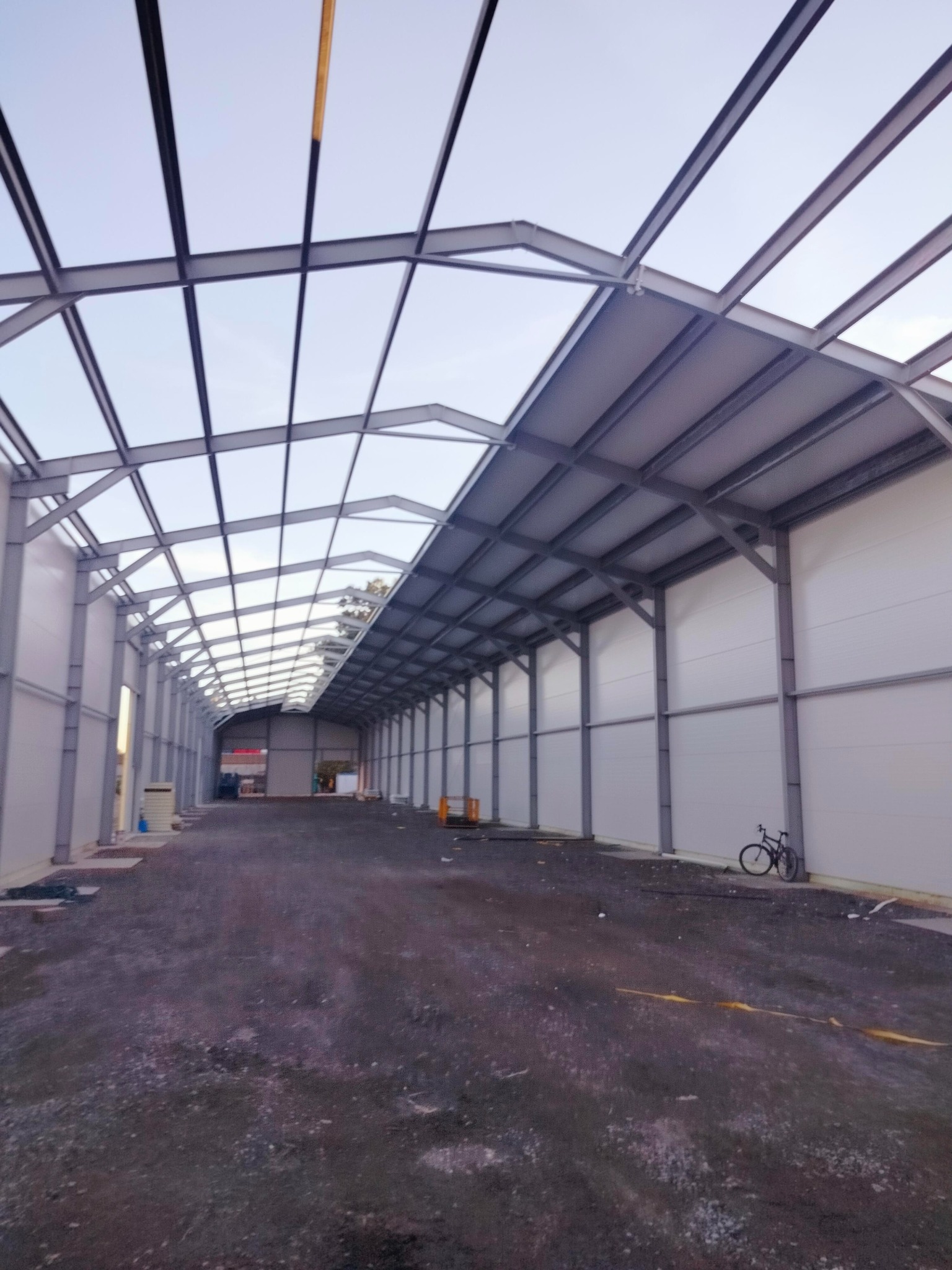 Wnętrze w trakcie budowy hali z widoczną stalową konstrukcją dachu i ścianami z płyt warstwowych, rower oparty o ścianę.