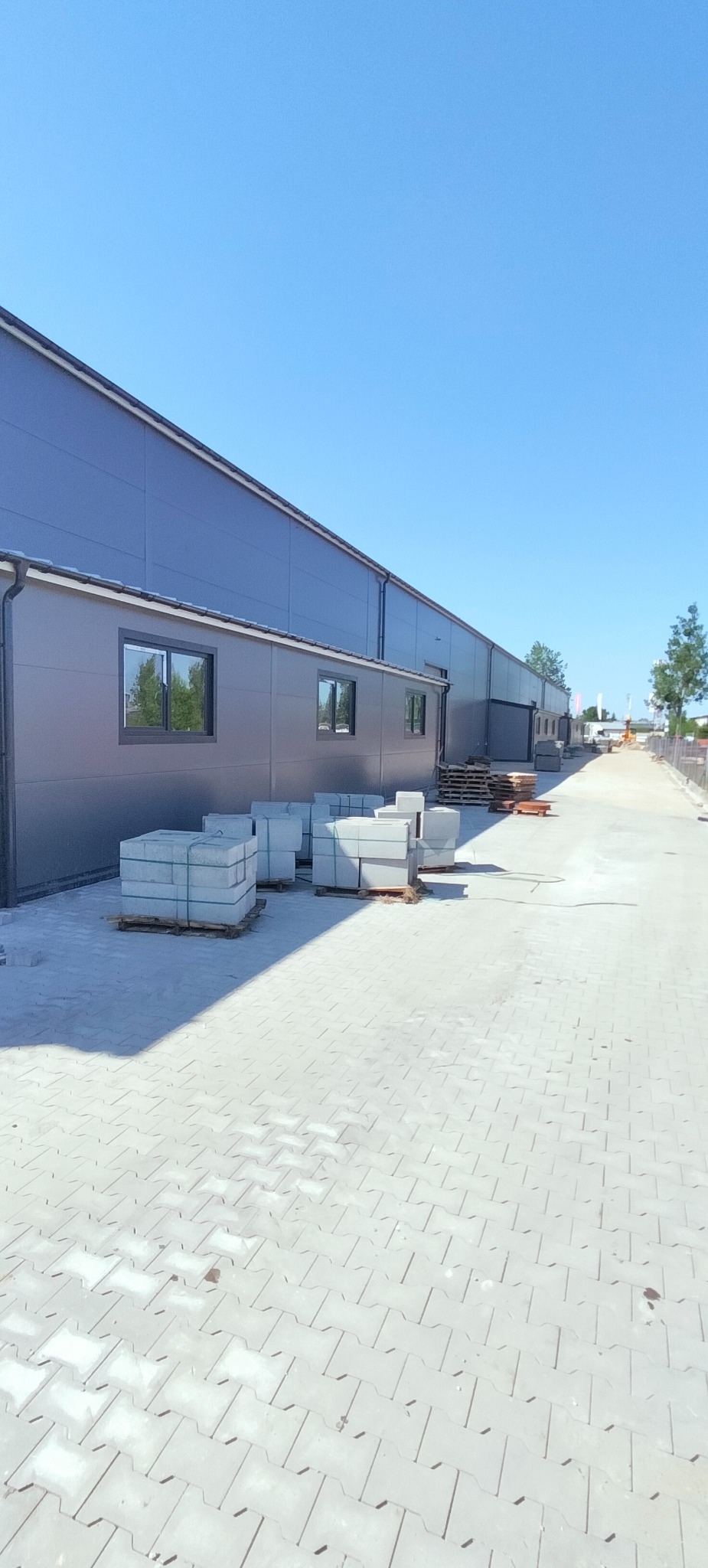 Gotowe bloki betonowe na paletach przed budynkiem hali z szarymi panelami, ułożone na kostce brukowej w słoneczny dzień.