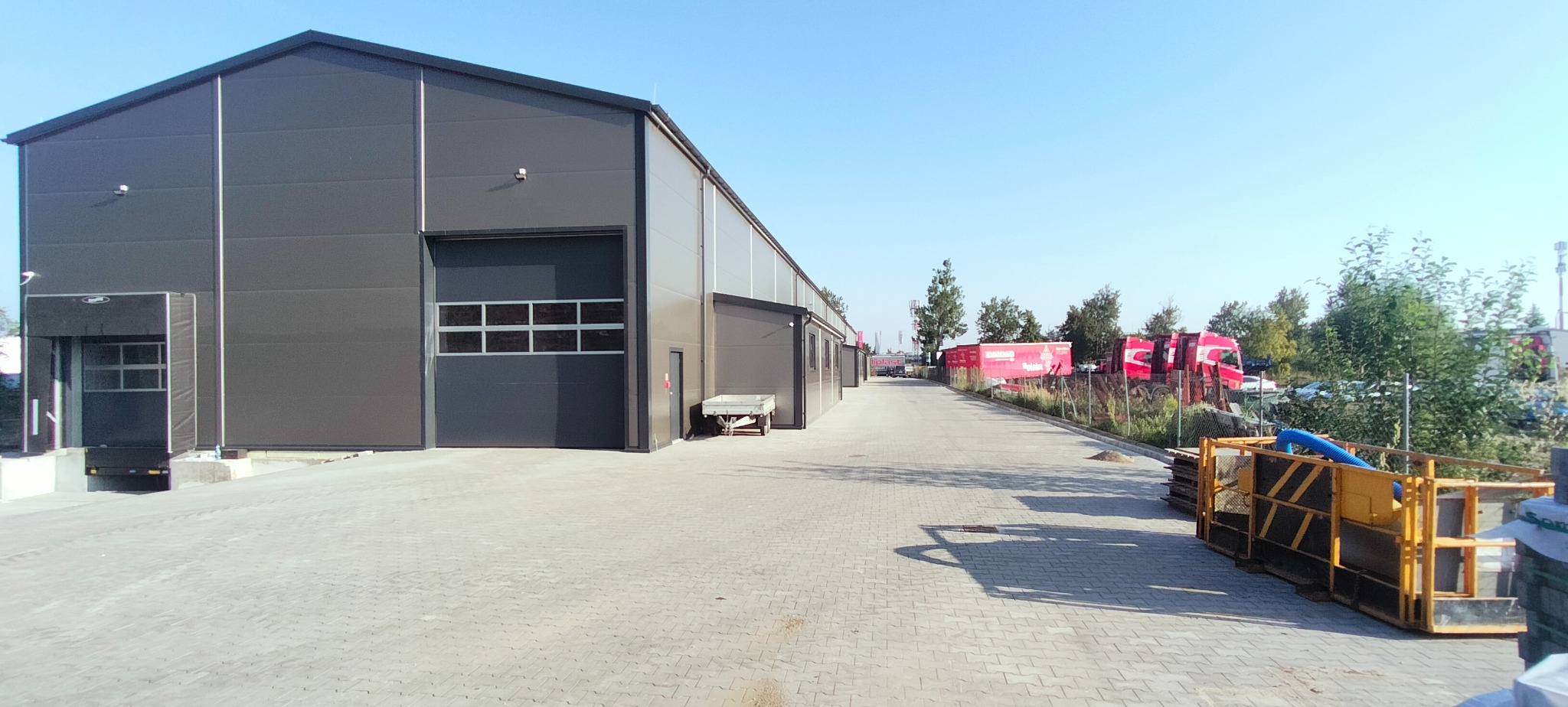 Nowoczesna hala magazynowa z szarej płyty warstwowej, widok z zewnątrz na plac z kostki brukowej i zaparkowane czerwone ciężarówki w tle.