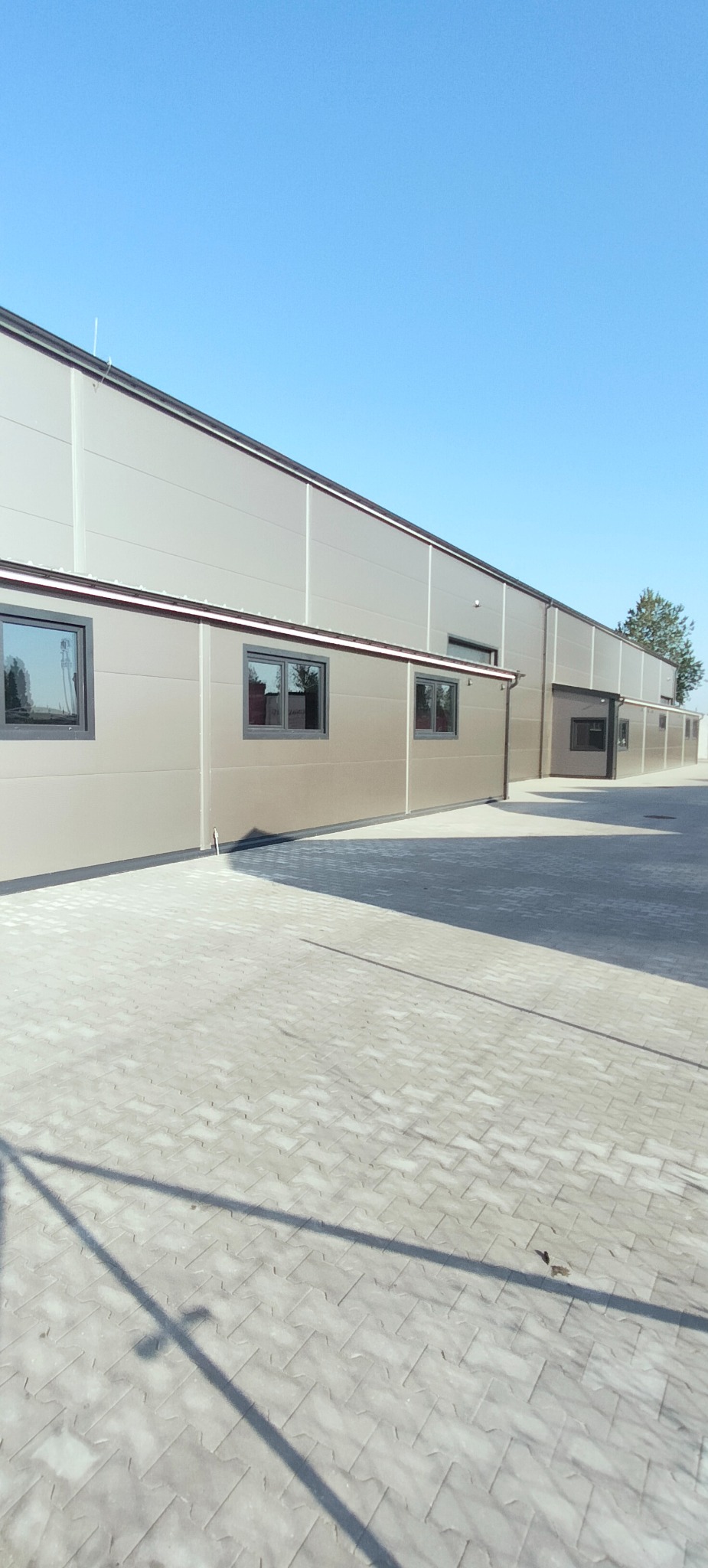 Nowoczesna hala przemysłowa z paneli warstwowych w odcieniach szarości, z widocznymi oknami i brukowaną nawierzchnią przed budynkiem w słoneczny dzień.