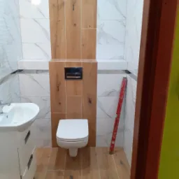 Wykończona łazienka z białymi marmurkowymi płytkami i drewnopodobnymi panelami, wisząca toaleta, umywalka z szafką oraz poziomica oparta o ścianę.