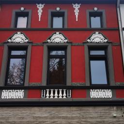 SandroSolution - Czerwona fasada kamienicy z ozdobnymi elementami sztukaterii wokół okien i pod nimi, widoczne detale architektoniczne i refleksy w szybach okiennych.