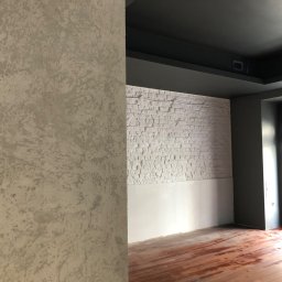 SandroSolution - Ściana z dekoracyjnym tynkiem w odcieniach szarości, kontrastująca z białą cegłą i ciemnoszarym sufitem w trakcie prac wykończeniowych w pomieszczeniu w Warszawie. Na podłodze widoczne ślady po...