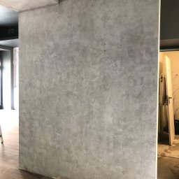 SandroSolution - Ściana z szarą, dekoracyjną strukturą tynku w trakcie prac wykończeniowych, widoczne zabrudzenia na podłodze i fragment zabezpieczenia podłoża.