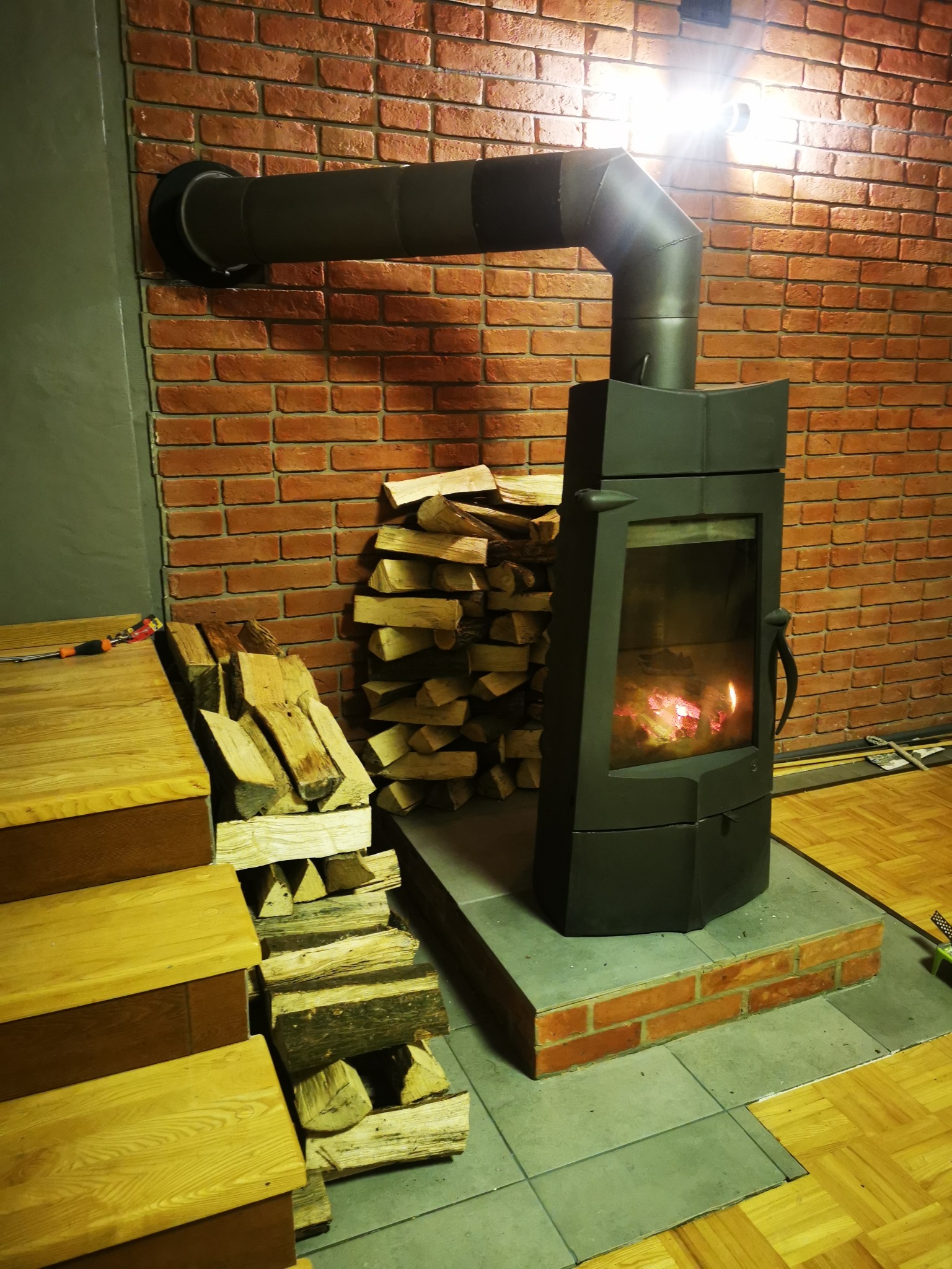 Ciemny piec wolnostojący z palącym się ogniem, z rurą odprowadzającą dym, ustawiony na szarych płytkach z ceglaną obwódką, obok ułożone drewno na opał, ściana z cegły.