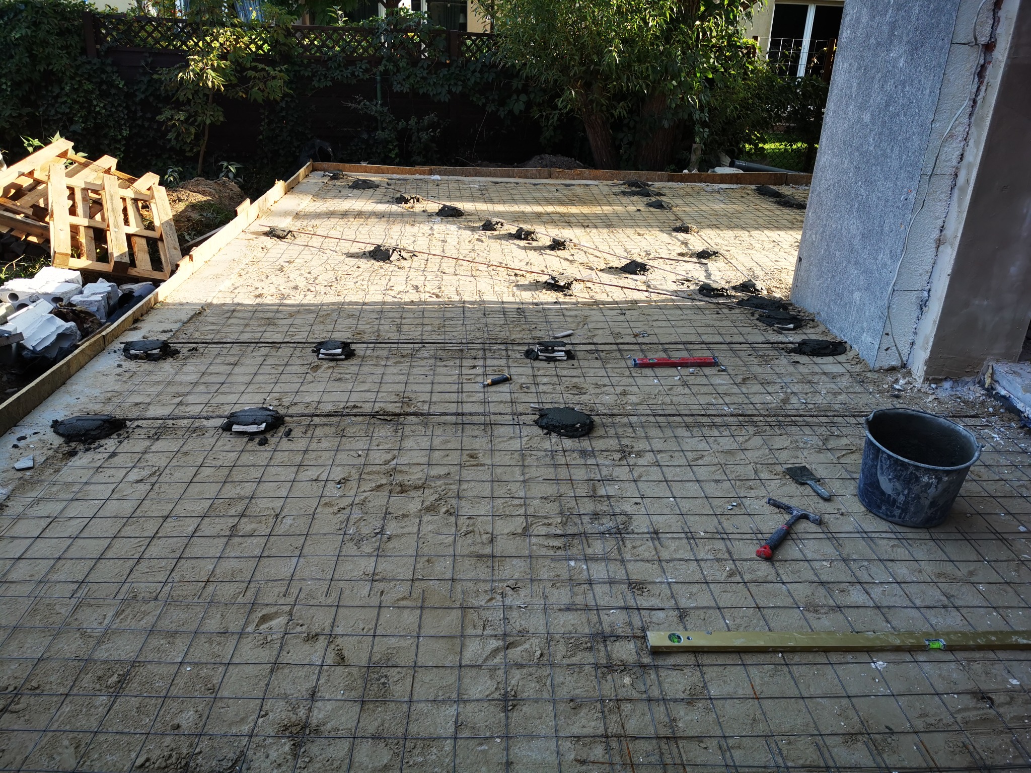 Przygotowanie podłoża pod taras: siatka zbrojeniowa, poziomica, wiadro i narzędzia na piasku, widoczne palety i roślinność w tle.