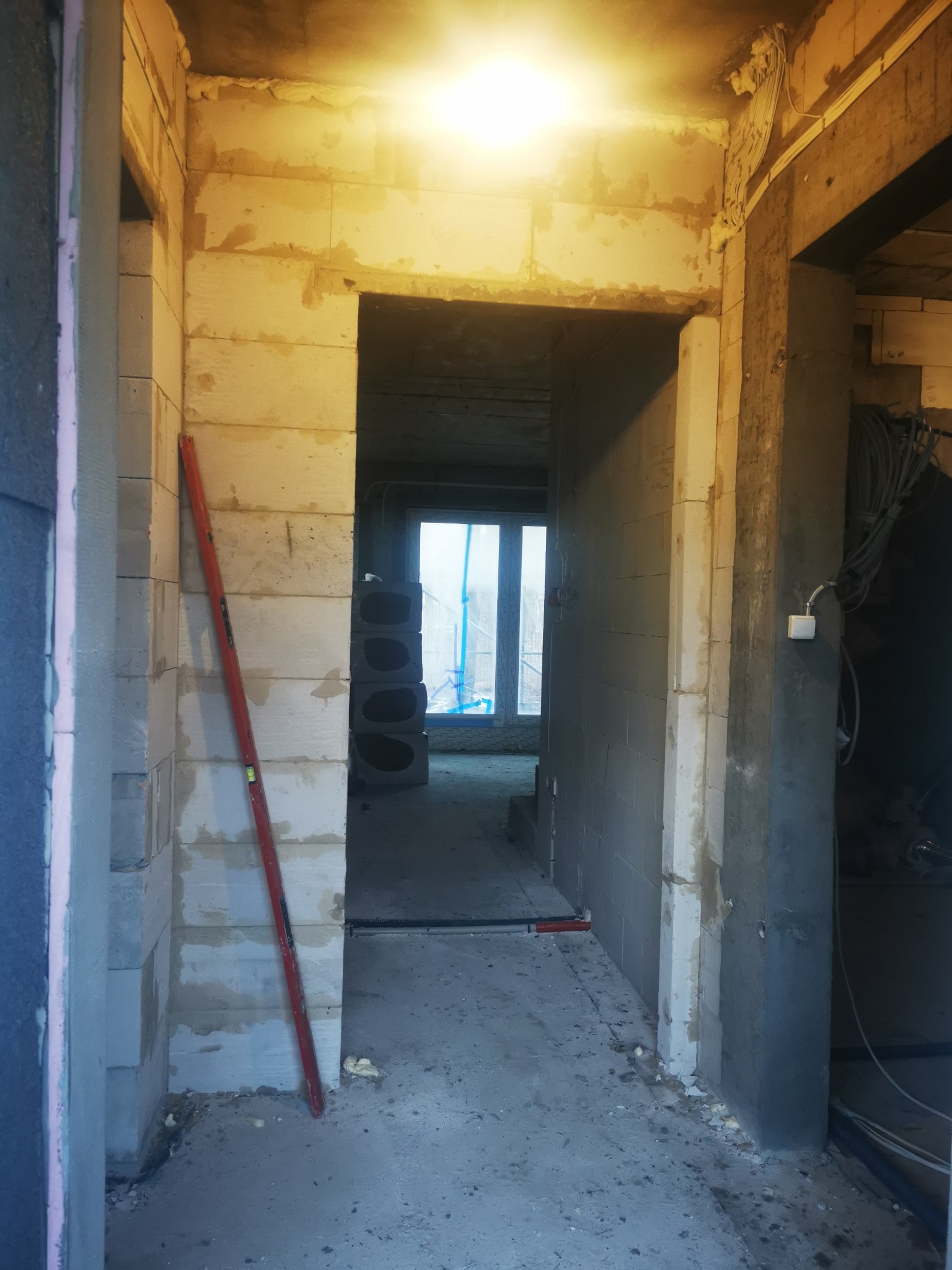 Wnętrze w trakcie prac murarskich: surowe ściany z betonu komórkowego, poziomica oparta o ścianę, widoczne przewody elektryczne pod sufitem, w tle okno z widokiem na zewnątrz.