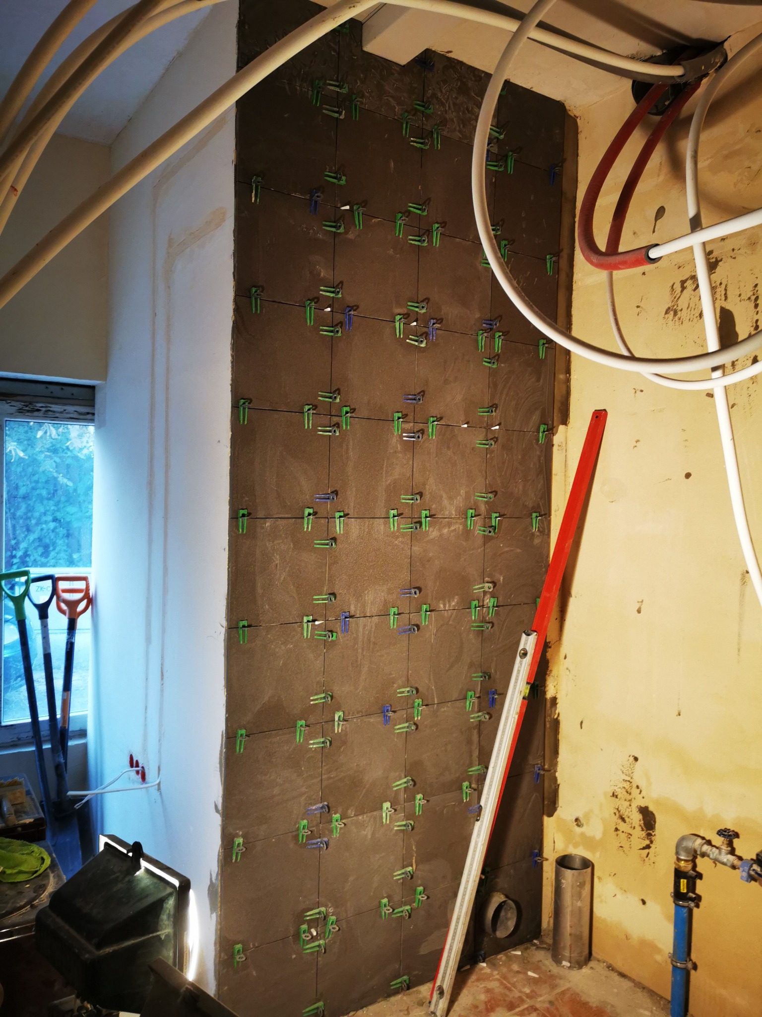 Układanie ciemnych płytek na ścianie z użyciem poziomicy i klipsów dystansowych, widoczne instalacje hydrauliczne i narzędzia w tle.