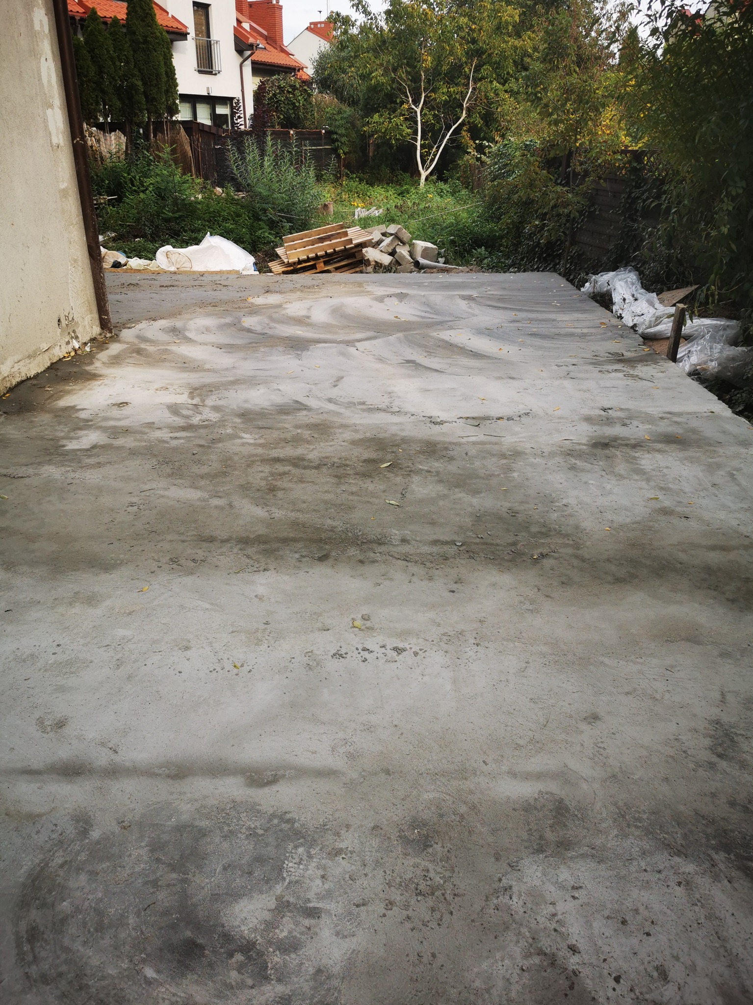Świeżo wylana, jeszcze mokra betonowa wylewka na zewnątrz budynku, widoczne ślady po zacieraniu, w tle palety i materiały budowlane przy ogrodzeniu.