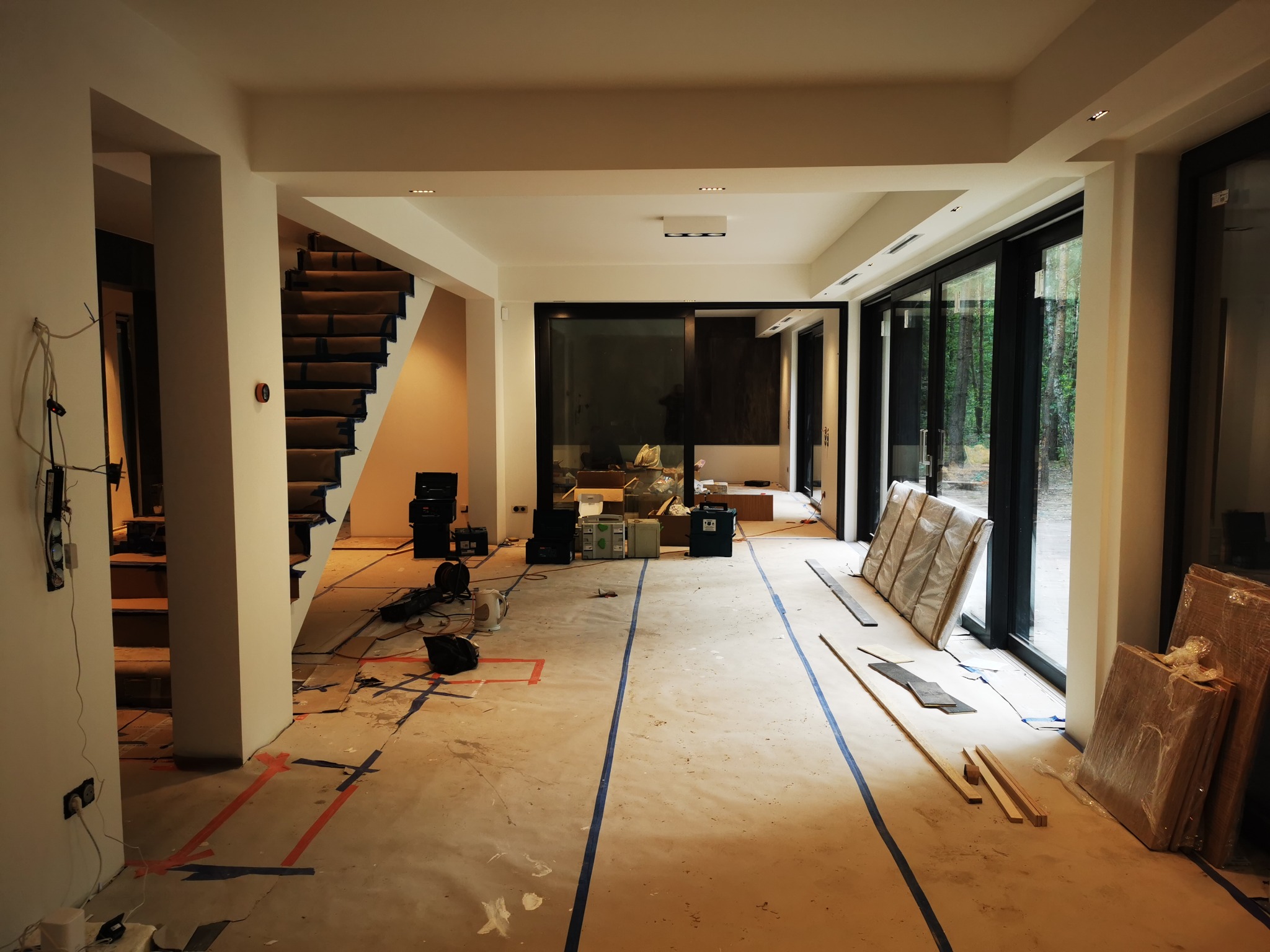 Salon w trakcie prac przygotowawczych do remontu: podłoga z taśmami wyznaczającymi obszary, zabezpieczone schody, okna przesuwne z widokiem na las, sprzęt budowlany w tle.