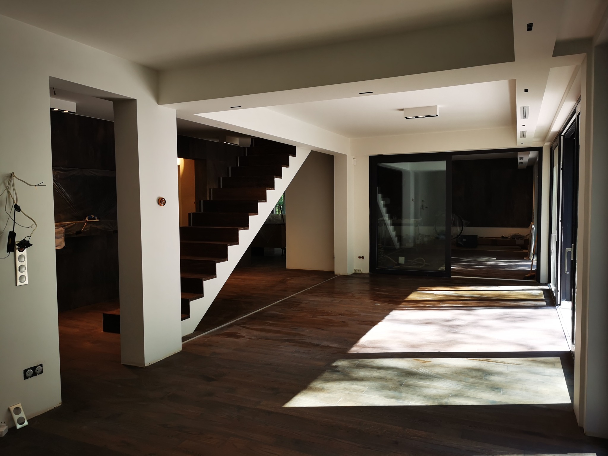 Wnętrze domu w trakcie wykończenia: drewniane schody, parkiet, duże przesuwne okna, widoczne niedokończone instalacje elektryczne przy ścianie.