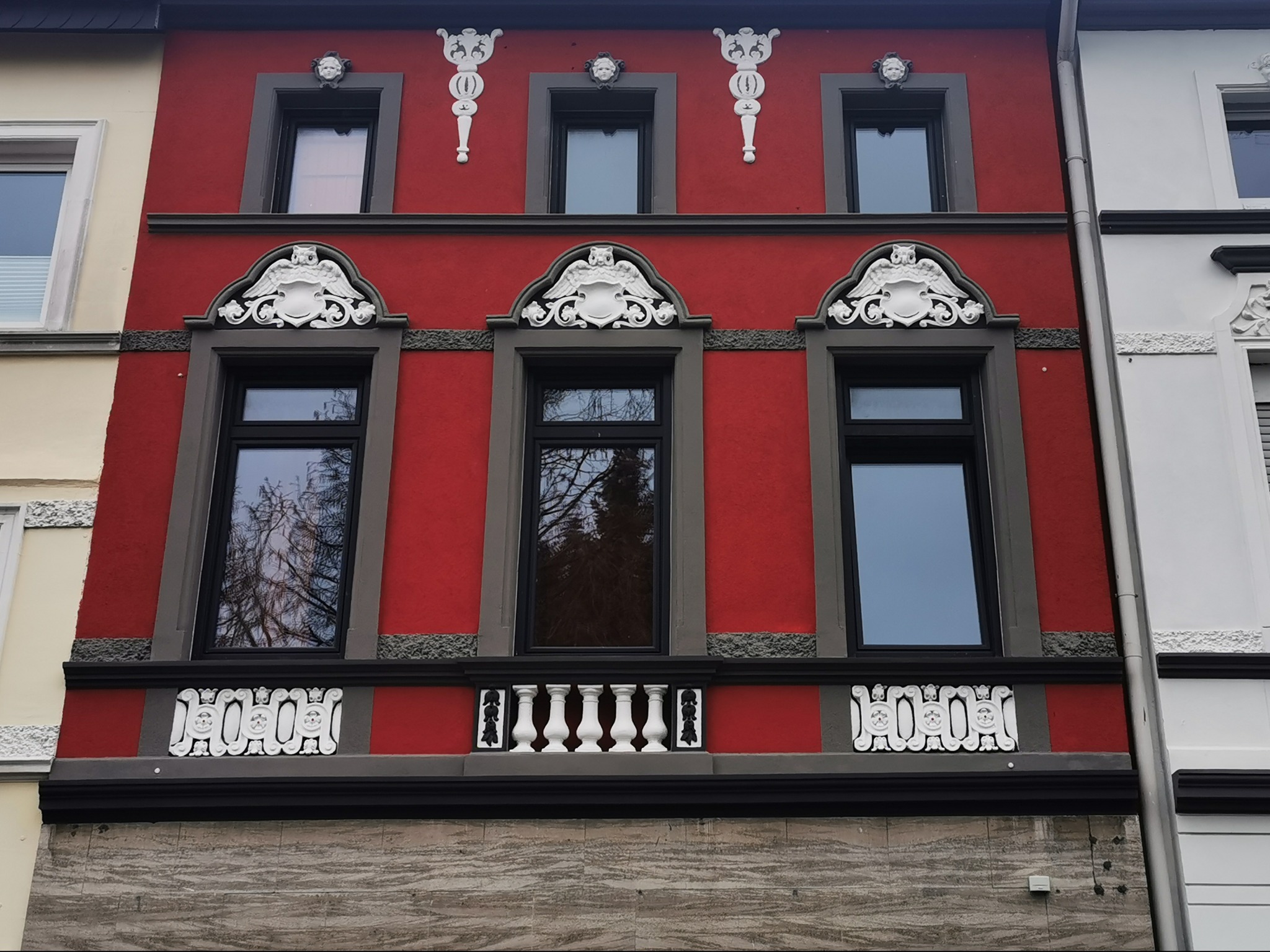 Czerwona fasada kamienicy z ozdobnymi elementami sztukaterii wokół okien i pod nimi, widoczne detale architektoniczne i refleksy w szybach okiennych.