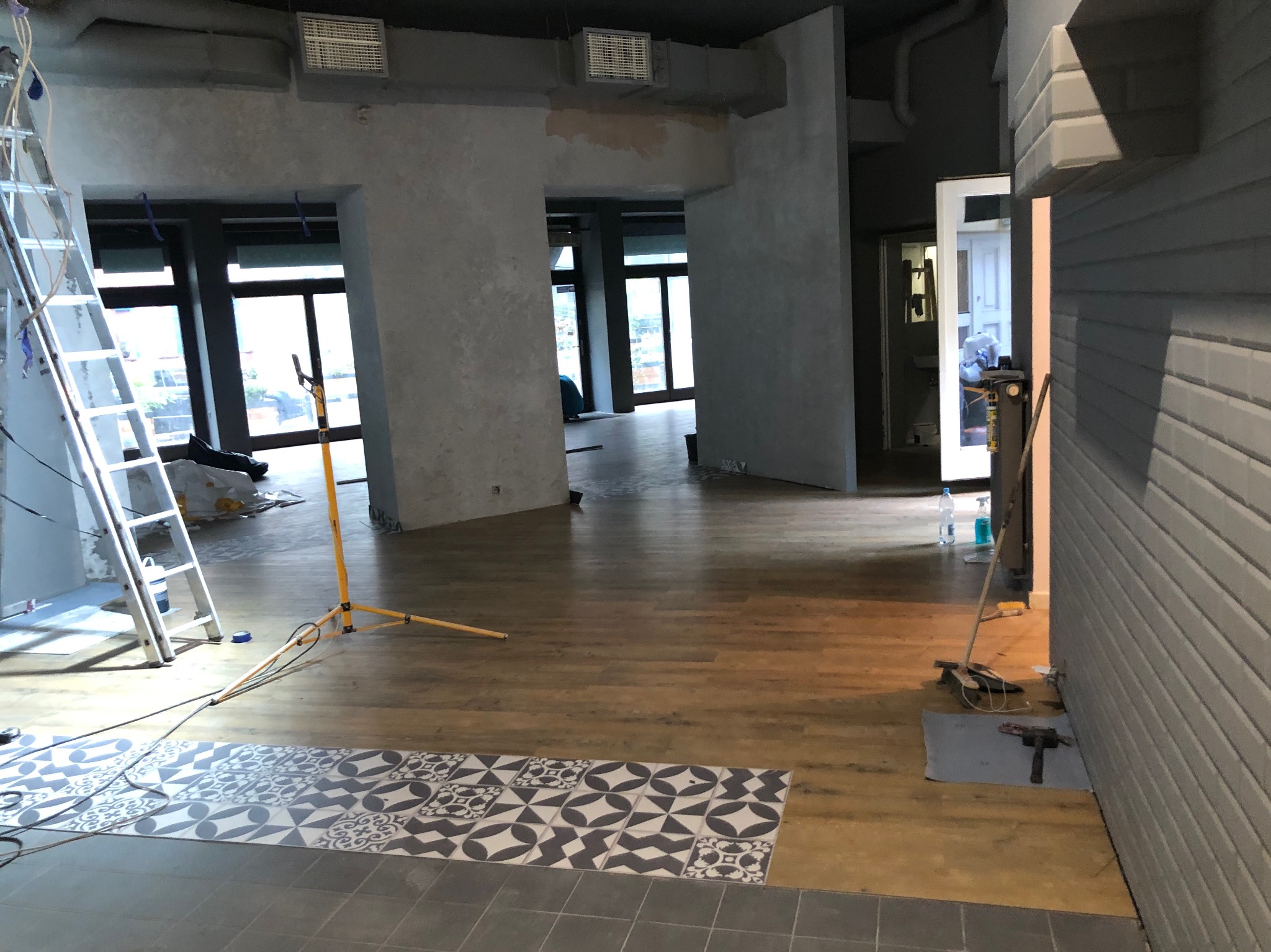 Wnętrze biura w trakcie prac wykończeniowych: częściowo położona podłoga z płytek o wzorze geometrycznym, widoczna drabina, lampa budowlana, ściany w surowym stanie, przygotowane do malowania.