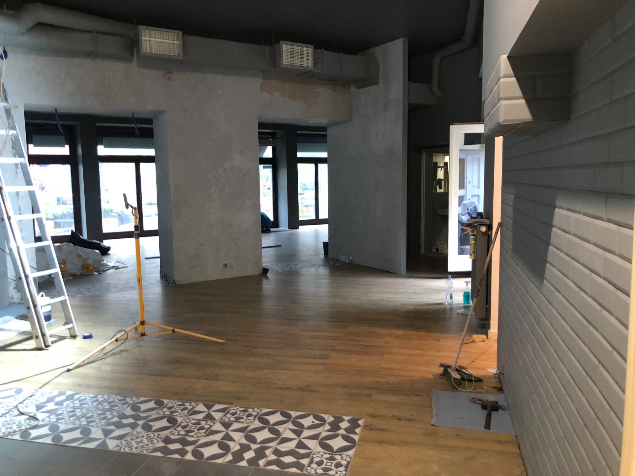 Wnętrze biura w trakcie prac wykończeniowych: surowy beton na ścianach, drewniana podłoga, fragment wzorzystej płytki, drabina, lampa budowlana, widoczne ślady szpachlowania i malowania.