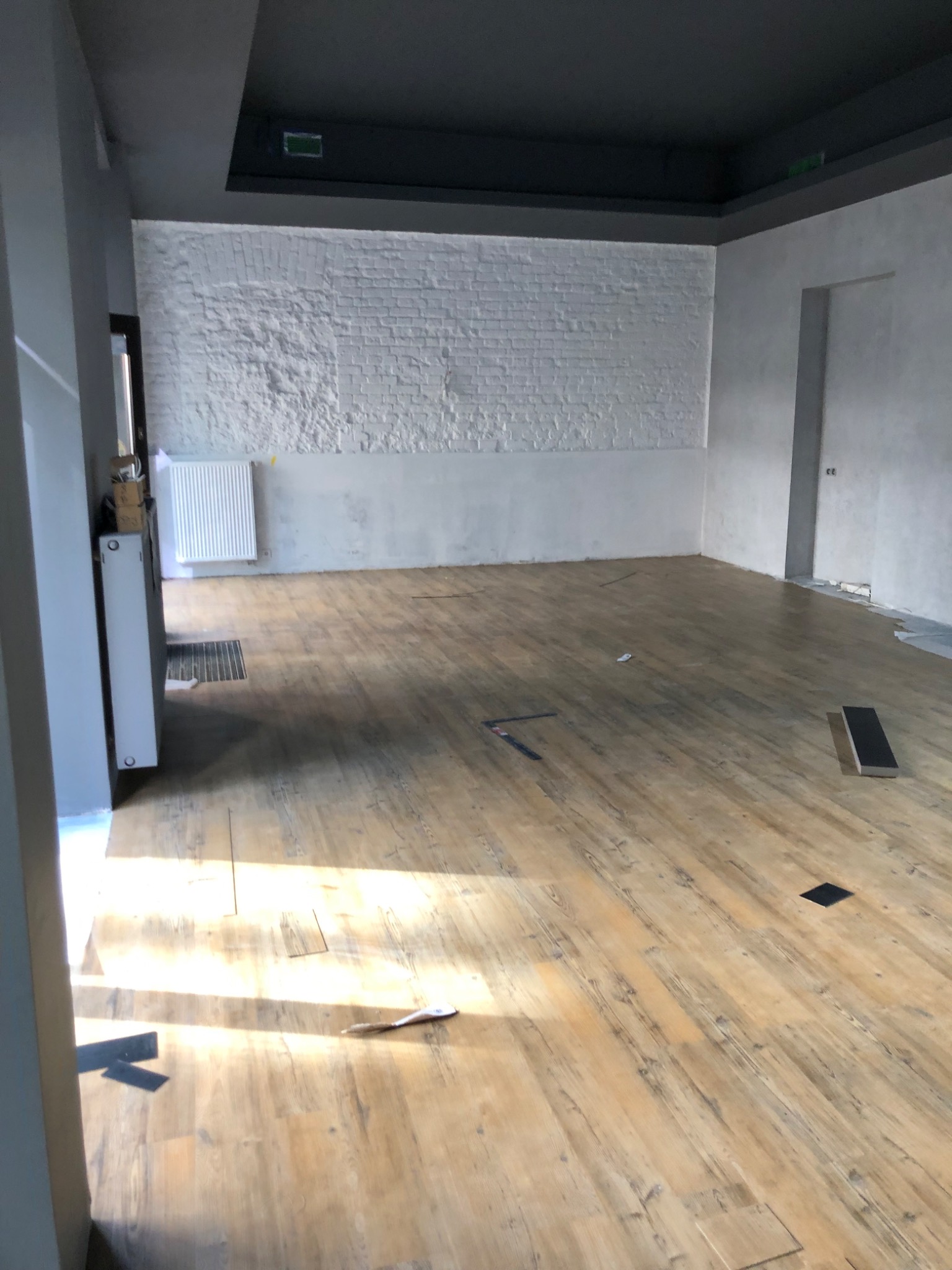 Wnętrze pustego pomieszczenia z drewnianą podłogą, białą ceglaną ścianą i szarym sufitem, w trakcie przygotowań do dalszych prac wykończeniowych, widoczny grzejnik oraz fragmenty niezabezpieczonej...