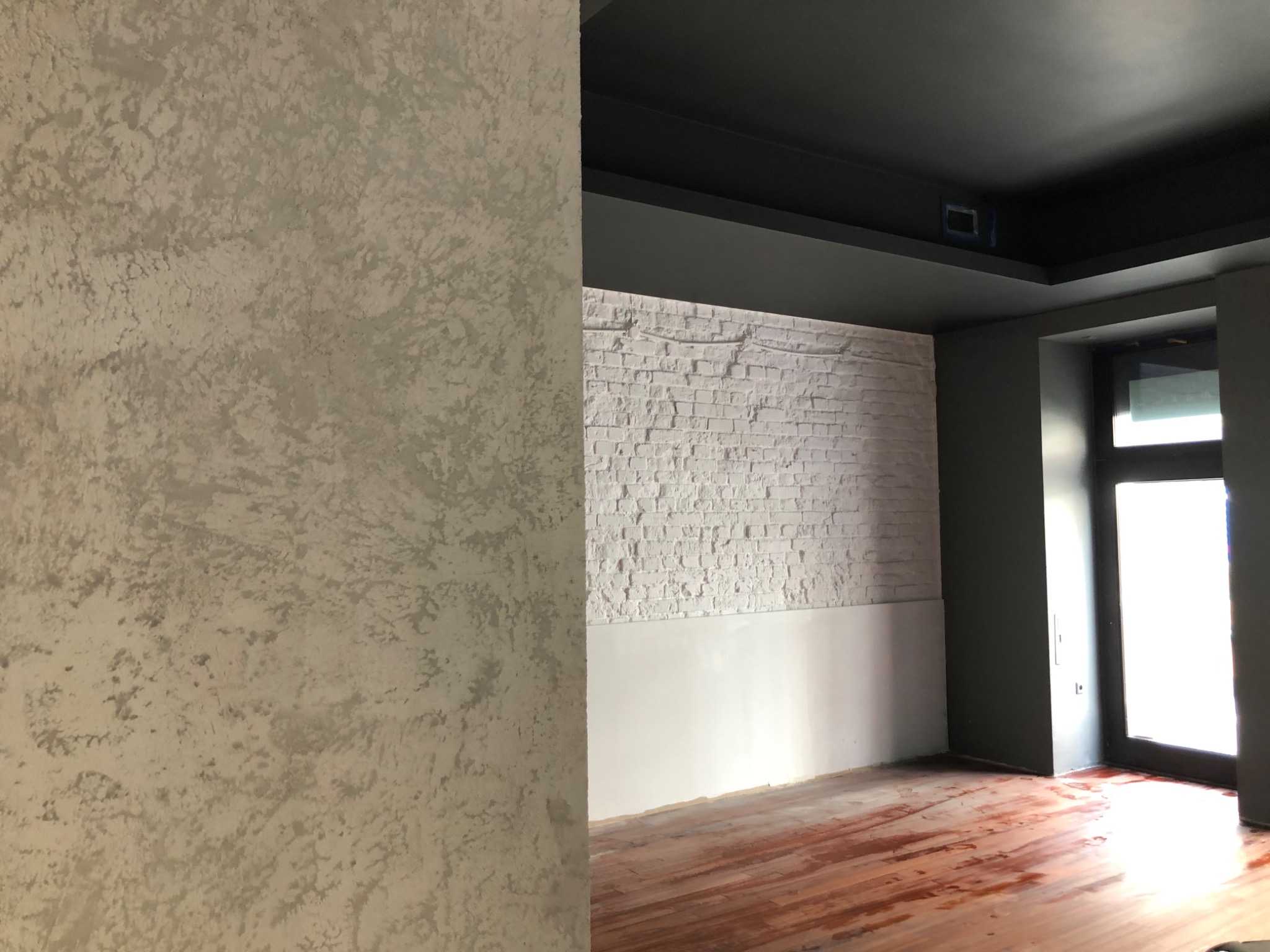 Ściana z dekoracyjnym tynkiem w odcieniach szarości, kontrastująca z białą cegłą i ciemnoszarym sufitem w trakcie prac wykończeniowych w pomieszczeniu w Warszawie. Na podłodze widoczne ślady po...