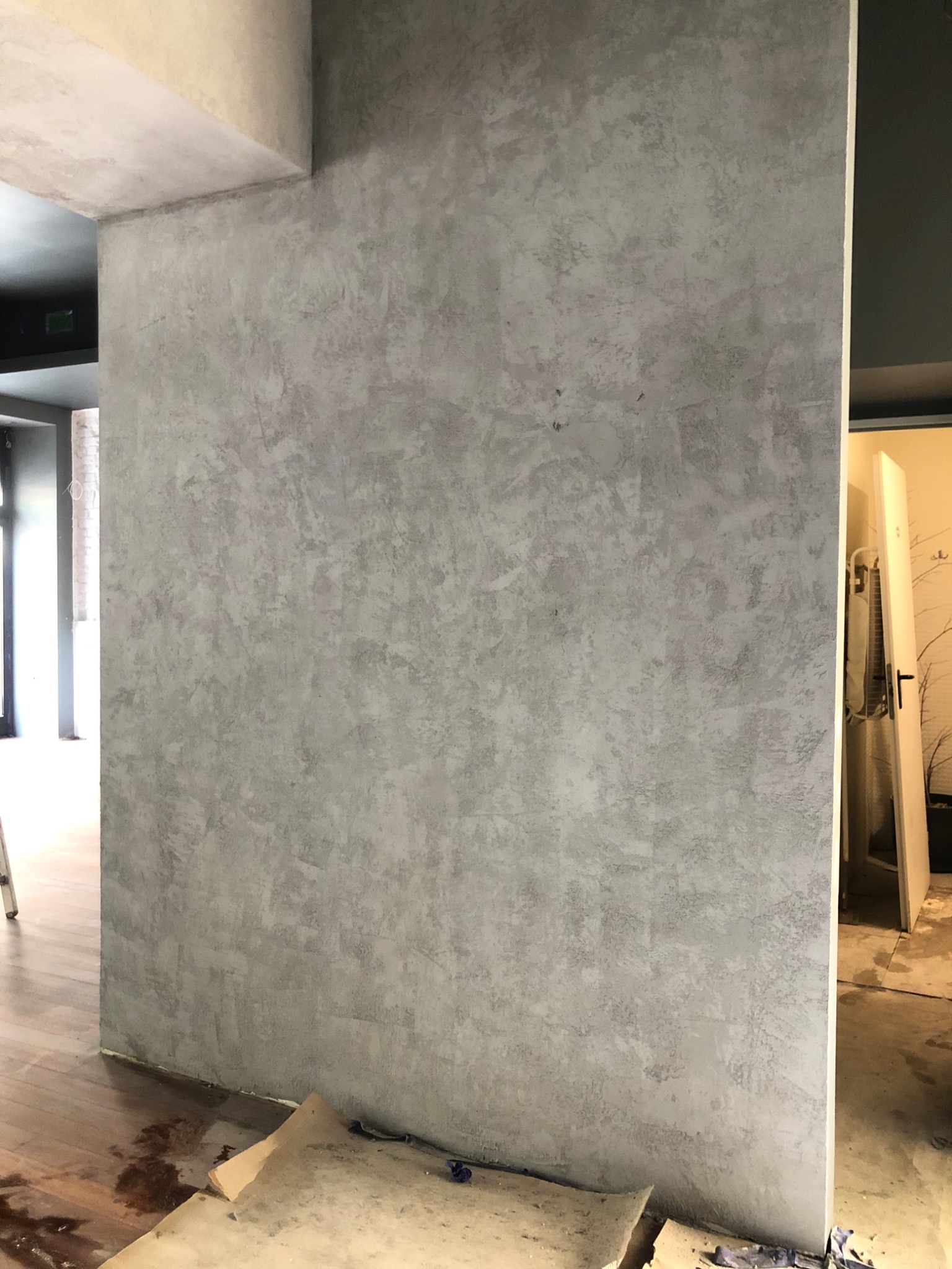 Ściana z szarą, dekoracyjną strukturą tynku w trakcie prac wykończeniowych, widoczne zabrudzenia na podłodze i fragment zabezpieczenia podłoża.