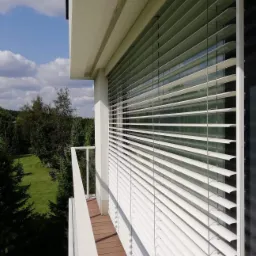 Białe żaluzje fasadowe zamontowane na balkonie nowoczesnego domu z widokiem na zieleń ogrodu.