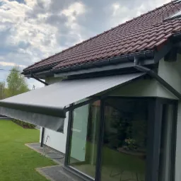 Szara markiza tarasowa zamontowana nad dużym oknem przesuwnym, widok z zewnątrz, trawnik przed domem.