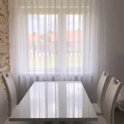 Jadalnia z białym, lakierowanym stołem i krzesłami na tle okna z delikatnymi, białymi zasłonami, fragment ściany z dekoracyjną cegłą.