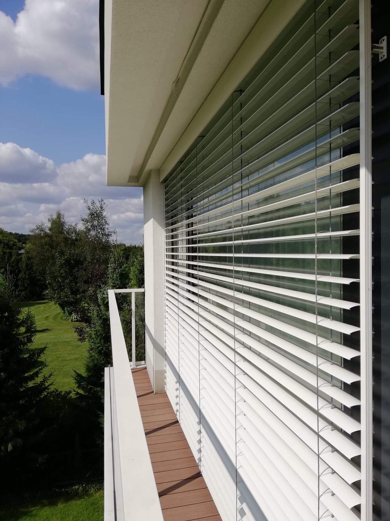 Białe żaluzje fasadowe zamontowane na balkonie nowoczesnego domu z widokiem na zieleń ogrodu.