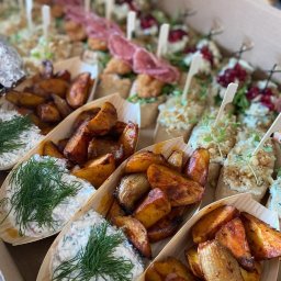 Pinchos_wroclaw - Aromatyczne pieczone ziemniaki z sosem ziołowym w drewnianych łódeczkach, obok kanapeczki z wędliną i owocami, ułożone w kartonowym pudełku.