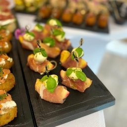 Pinchos_wroclaw - Elegancko podane przekąski: brzoskwinie owinięte szynką parmeńską z mini mozzarellą i bazylią, na czarnych kamiennych deskach, w tle inne finger foody.