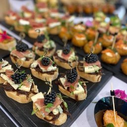 Pinchos_wroclaw - Elegancko podane kanapeczki z brioletami, szynką parmeńską i serem pleśniowym, udekorowane jeżynami i świeżymi ziołami, ustawione na czarnej tacy obok miseczek z krewetkami.
