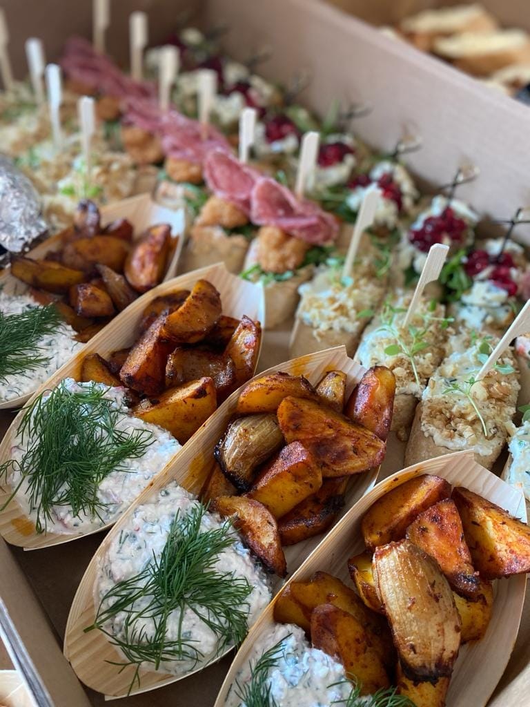 Aromatyczne pieczone ziemniaki z sosem ziołowym w drewnianych łódeczkach, obok kanapeczki z wędliną i owocami, ułożone w kartonowym pudełku.