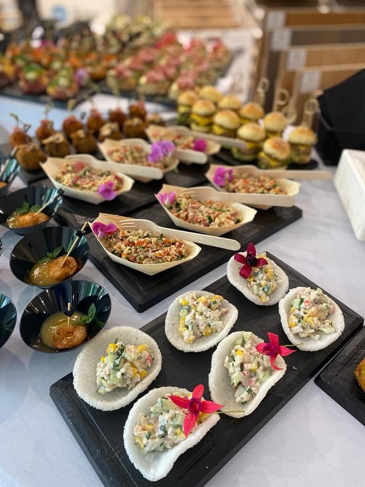 Różnorodne przekąski na czarnych deskach i białym obrusie: mini burgery, sałatki w łódeczkach z drewna i ryżu, kulki mięsne, dekorowane kwiatami.