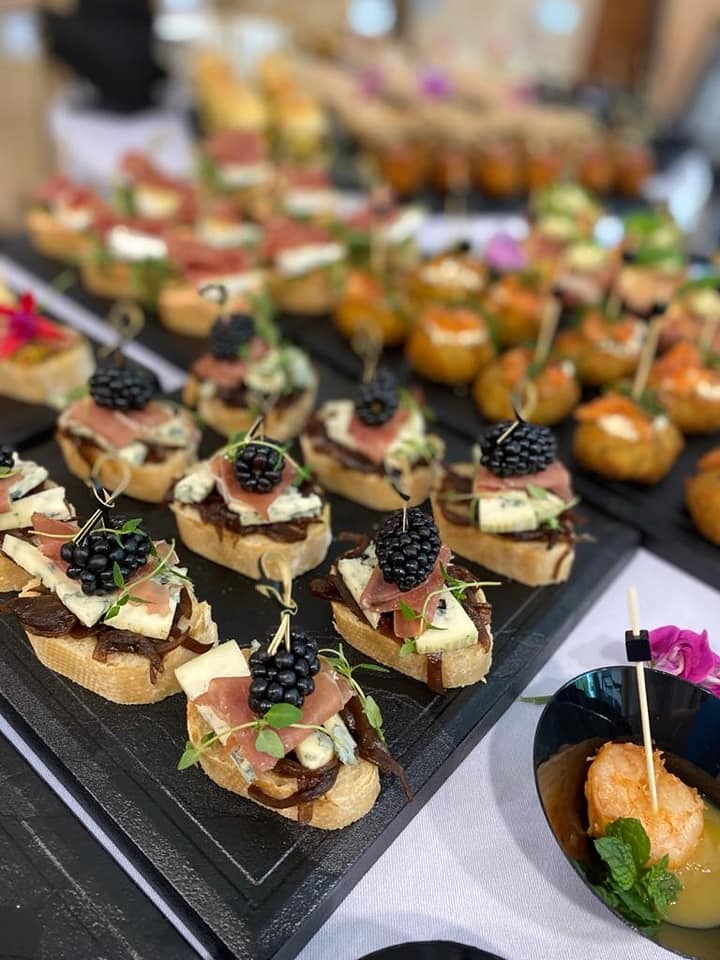 Elegancko podane kanapeczki z brioletami, szynką parmeńską i serem pleśniowym, udekorowane jeżynami i świeżymi ziołami, ustawione na czarnej tacy obok miseczek z krewetkami.