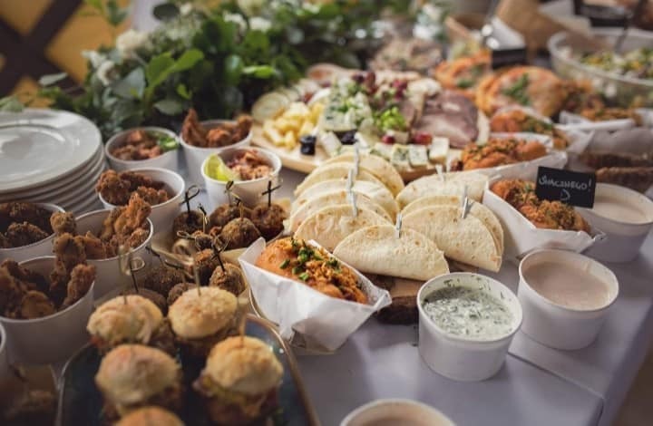 Bogato zastawiony stół cateringowy z mini burgerami, tortillami z wykałaczkami, kulkami mięsnymi na patykach, smażonym kurczakiem w kubełkach, serami i wędlinami na drewnianej desce, oraz sosami...