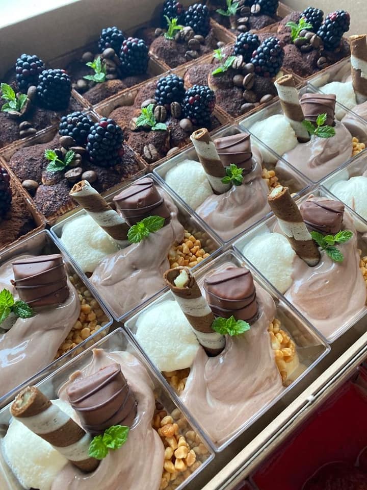 Zestaw deserów w przezroczystych pojemnikach: tiramisu z jeżynami i miętą oraz krem czekoladowy z waflami, bitą śmietaną i orzechami, ułożonych w kartonowym pudełku.