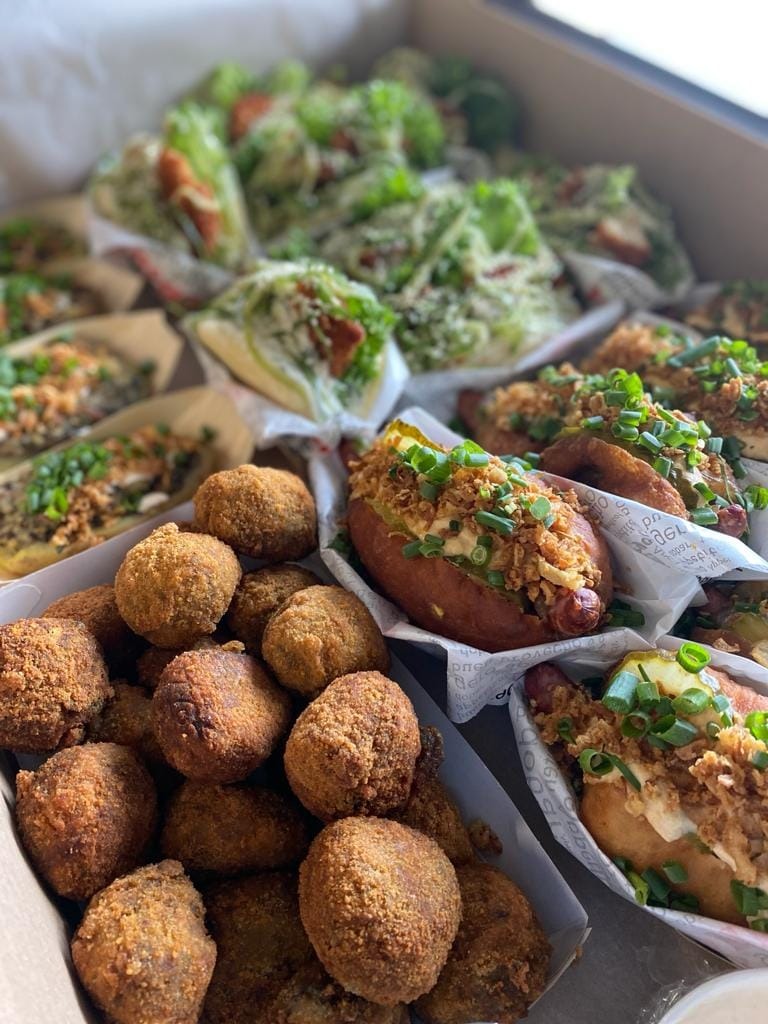 Różnorodne przekąski w kartonowym pudełku: smażone kulki, hot dogi z prażoną cebulką i szczypiorkiem oraz sałatki w bułkach.