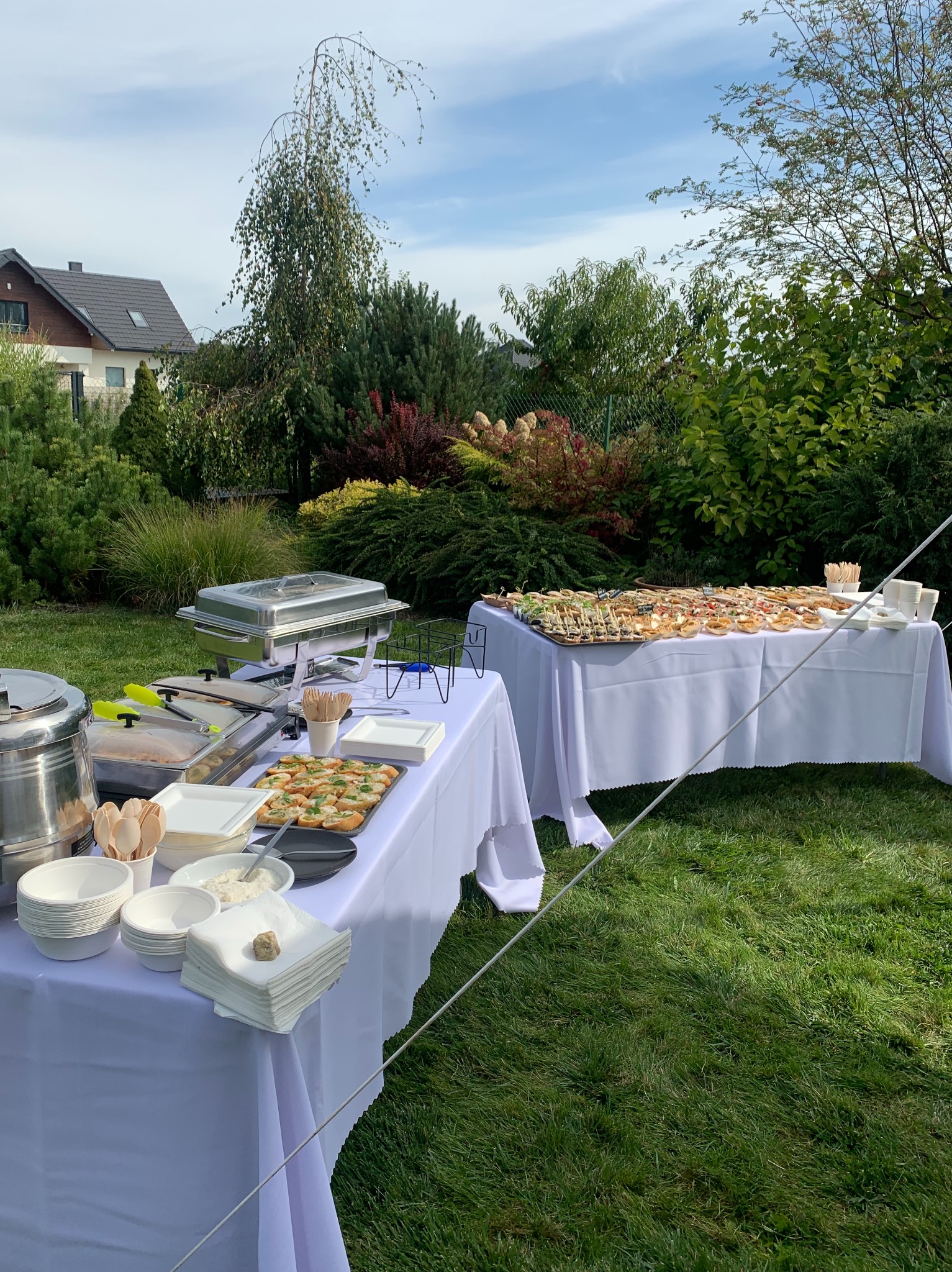 Stół z białym obrusem na trawie, bufet z przekąskami, podgrzewacze, naczynia i serwetki, w tle ogród z domem.