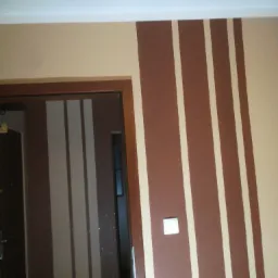 Ściana z dekoracyjnym malowaniem w pionowe pasy w odcieniach beżu i brązu, widoczny fragment futryny drzwiowej i włącznik światła.