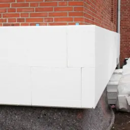 Fragment ocieplonej ściany budynku z czerwonej cegły, widoczne płyty styropianowe z niebieskimi kołkami mocującymi, obok stos zapakowanych płyt styropianowych.